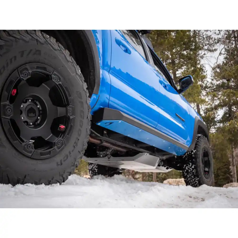 Angled view of Blue Tacoma with black tires on a snowy surface with mounted skid plates